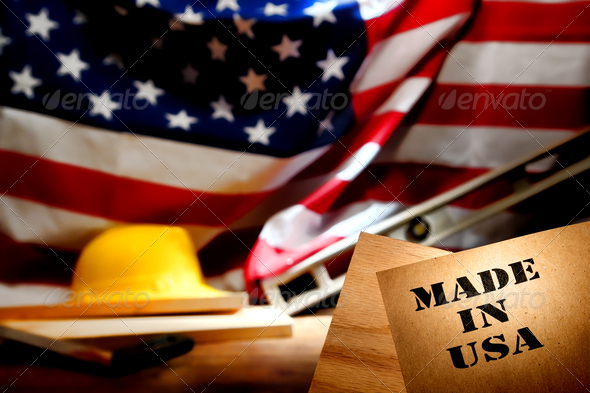 Made in USA stencil stamp logo on brown recycled paper over a carpentry wood board at an American construction work site in the United States with patriotic US flag and trade working tools in background