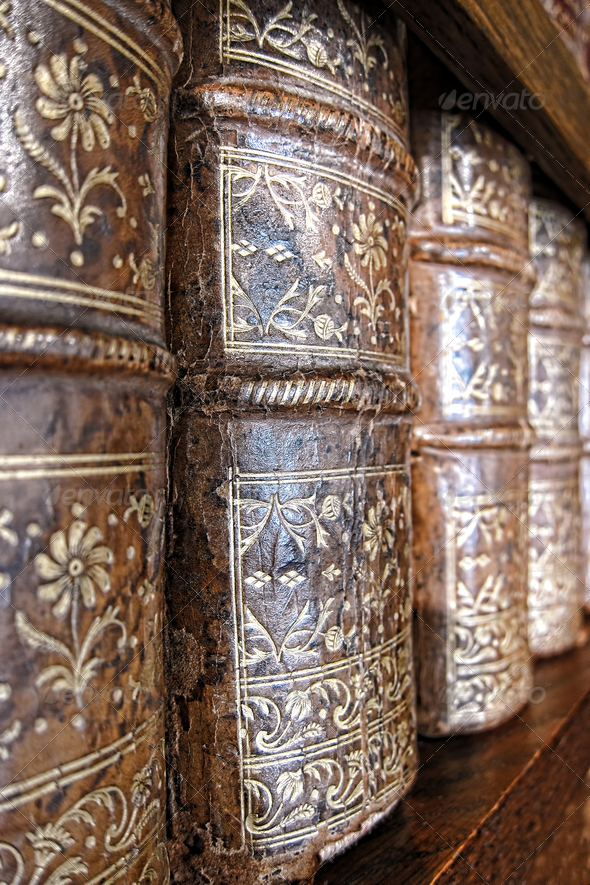 Old and worn leather cover bound books spine with aged gold leaf embossing on an antique wood library bookcase shelf