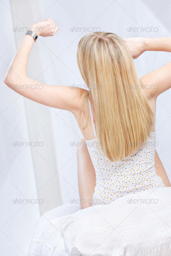 Back of a long blond hair woman sitting on bed, stretching arms and looking at the clock