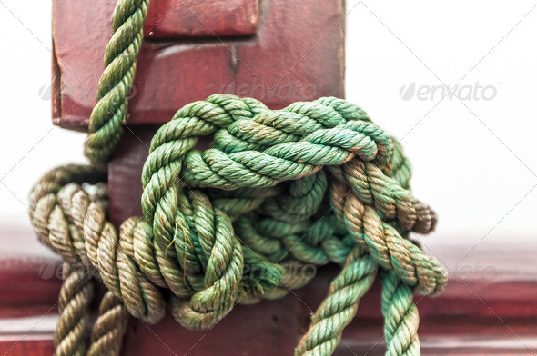 Marine rope tied into knot in foreground, white background. Close-up of nautical equipment for mooring. Focus on detail of ship. Green twisted rope wrapped around red wooden deck. Sea travel.