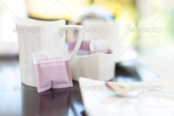 White clean dishware on shining table in cafe with focus on mug and packet of brown sugar. Blurry empty plate with spoon in foreground. Having lunch in cafe or restaurant. Coffee or tea break.