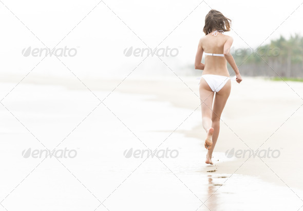 Pretty young woman in white swimsuit running on wet sand along ocean beach leaving footprints. View from back. Summer holidays and vacations at seaside. Relaxation at coast.