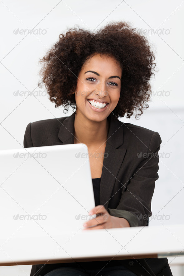 Happy African American businesswoman or student with an afro hairstyle sitting at a table with a laptop computer looking at the camera with a friendly smile