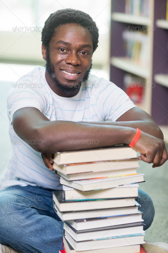 Full length portrait of smiling male student with stacked books sitting on floor at library