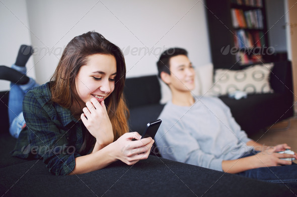 Pretty young woman lying on couch texting on mobile phone while young man playing video game in background at home. Mixed race teenage couple in living room indoors.